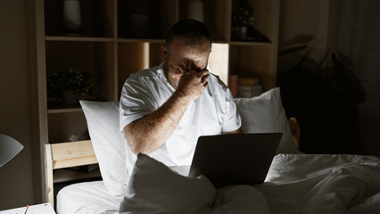 Exhausted caucasian man slumped over laptop while sitting on bedroom bed, stressed from overwork in...