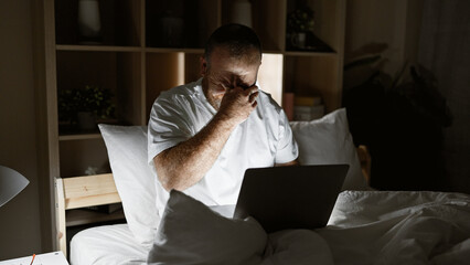 Exhausted caucasian man slumped over laptop while sitting on bedroom bed, stressed from overwork in dimly lit room