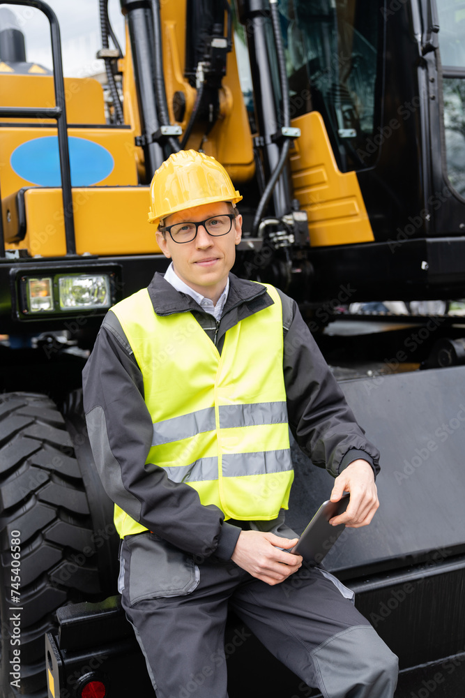 Poster engineer in a helmet with a digital tablet stands next to construction excavators..