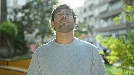 Hispanic young man with closed eyes, enjoying a breathing meditation in a sunny city park, a symbol...