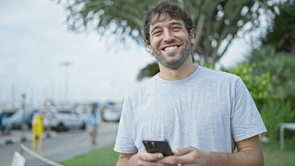 Fototapeta premium Happy young man, sporting a beard and blond hair, enjoying his time outdoor in the city, confidently smiling while typing a message on his smartphone screen