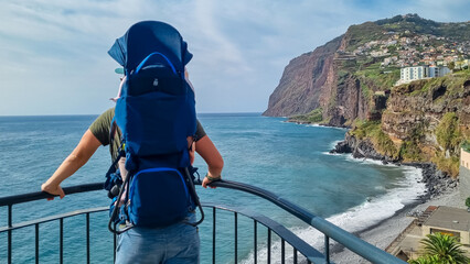 Tourist man wearing baby carrier looking at Praia de Vigario in Camara de Lobos on Madeira island, Portugal, Europe. Black stone beach in Atlantic Ocean. Colourful house on steep hills and cliffs - obrazy, fototapety, plakaty