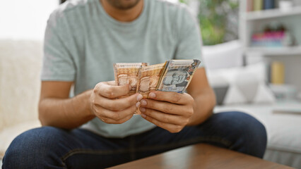 Hispanic young man in living room, sitting on sofa, indulged in counting his wealth - peruvian soles banknotes, mastering home finance. making banking and investment decisions indoors.