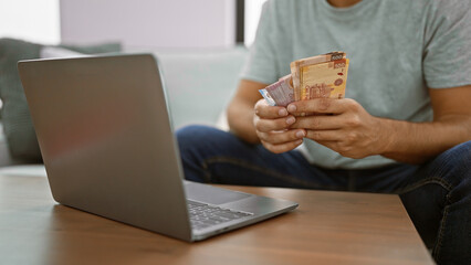 Young man sitting at home, banking online on his laptop, holding wads of rich mexican peso banknotes on his apartment's sofa. - obrazy, fototapety, plakaty
