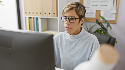 Hispanic woman with short blonde hair and glasses working attentively at a computer in a modern...