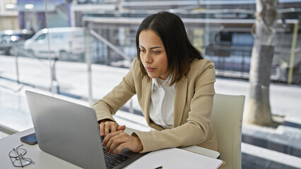 Focused hispanic woman working on her laptop at a modern office desk, exemplifying professionalism and concentration.