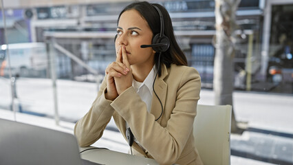 A contemplative hispanic woman in a headset sits at a desk against a blurred office backdrop.