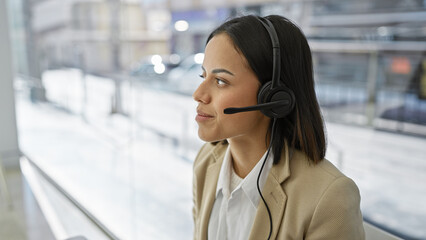 A poised young woman wearing a headset stands against a blurred indoor background, radiating...