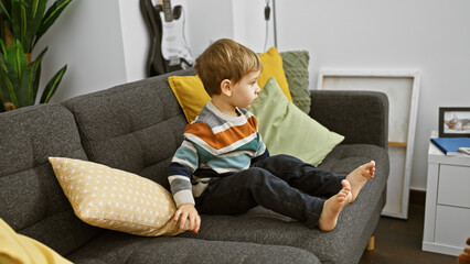 Blond toddler sitting calmly on a grey sofa in a modern living room, gazing thoughtfully.