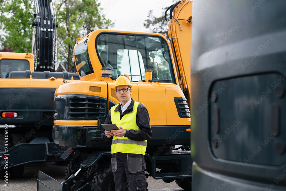 Poster Engineer in a helmet with a digital tablet stands next to construction excavators..