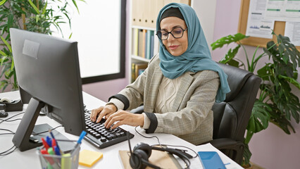A professional woman wearing a hijab working diligently at an office desk with a computer,...