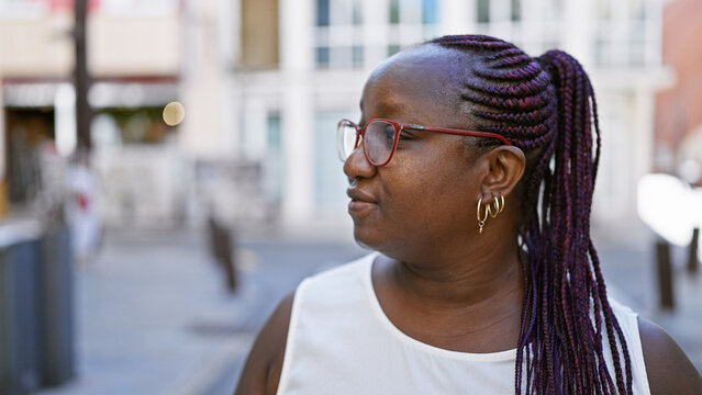 Cool plus size african american woman in glasses, with braids, casting a serious expression while looking to the side on a sunny urban street. - Powered by Adobe
