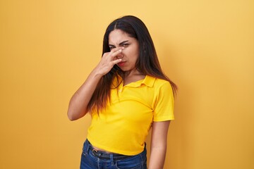 Young arab woman standing over yellow background smelling something stinky and disgusting,...