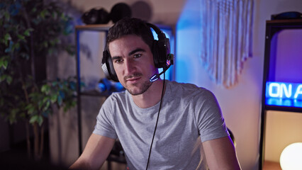 A young hispanic man wearing headphones in a dark indoor gaming room lit by neon lights, portraying...