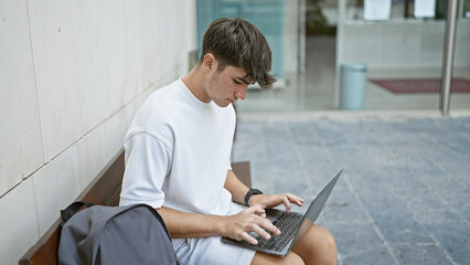 Cool young hispanic teenager guy, a relaxed student, concentrates on his laptop sitting on a city university bench, expressing his serious yet smart lifestyle outdoors.
