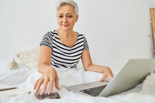 Attractive Gray Haired Elderly Businesswoman Working Remotely Right From Bedroom Sitting Bed With Portable Computer Using Calculator Managing Finances Having Confident Happy Expression