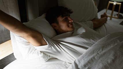 Happy young hispanic teenager stretching arms in comfort of his morning bed, waking up confidently...
