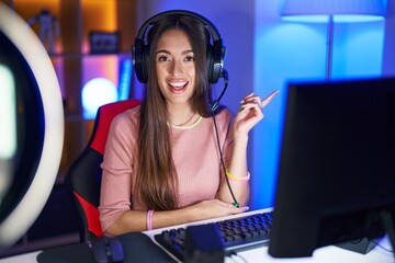 Young hispanic woman playing video games with a big smile on face, pointing with hand and finger to the side looking at the camera.