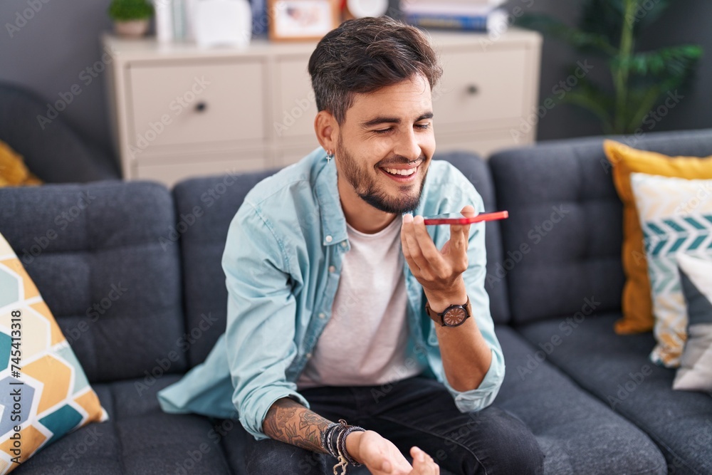 Sticker young hispanic man talking on the smartphone sitting on sofa at home