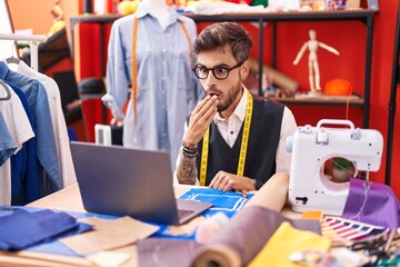 Young hispanic man with tattoos dressmaker designer using laptop at tailor room covering mouth with...