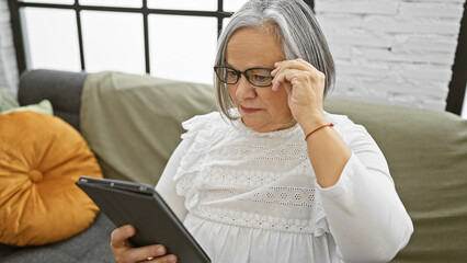 A senior woman with grey hair and glasses focused on reading a tablet indoors, conveying a sense of technology use in elderly.