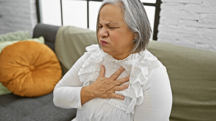 Fototapeta premium A senior woman experiencing chest pain while sitting on a couch in a well-lit living room.