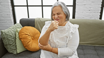 A senior woman grimacing in pain holds her elbow in a modern living room.