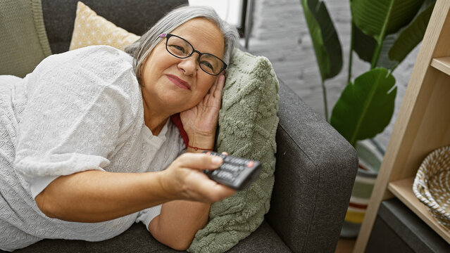 A smiling senior woman lounging on a couch at home with a remote control in hand, surrounded by cozy decor.
