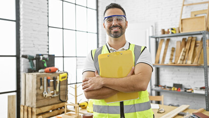 A smiling bearded hispanic man in a reflective vest with safety goggles holds a clipboard in a...
