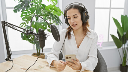 Hispanic woman podcasting indoors using microphone and smartphone, creating content in a modern...