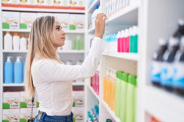 Poster Young blonde woman customer holding product of shelving at pharmacy © Krakenimages.com