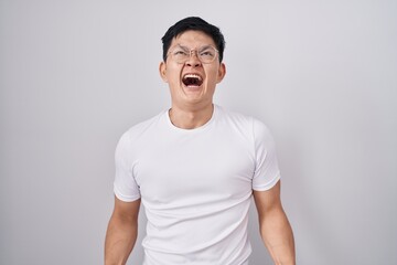 Young asian man standing over white background angry and mad screaming frustrated and furious, shouting with anger. rage and aggressive concept.