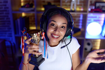 Young arab woman playing video games holding trophy celebrating achievement with happy smile and...