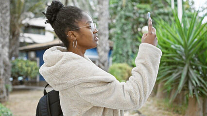 African american woman taking selfie outdoors in casual attire with smartphone, urban garden...