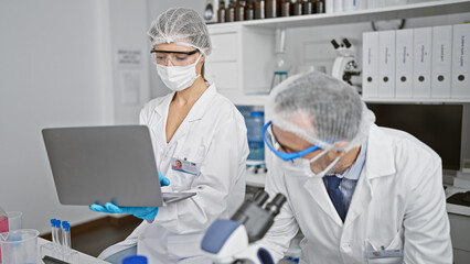 Naklejka premium Together in the lab, two scientists sporting medical masks, zooming in with their microscope while working on their laptop amid covid19 research.