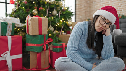 Obraz na płótnie Canvas Young hispanic woman sitting on the floor wearing christmas hat looking bored at home