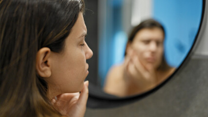 Fototapeta premium Young beautiful hispanic woman looking for pimples at the mirror in the bathroom