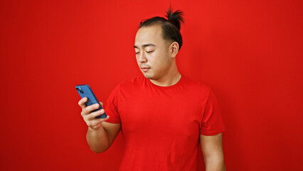 Handsome young chinese man, in relaxed concentration, uses his phone, tapping into the digital world over an isolated red background