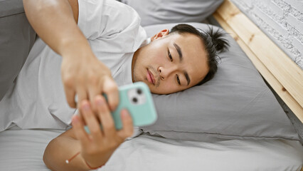 Obraz na płótnie Canvas Handsome young chinese man, in relaxed fashion, lying comfortably in bed at home using smartphone in bedroom, immersed in a serious online world