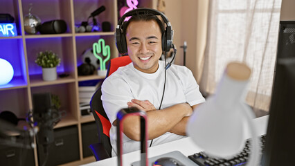 Smiling young chinese streamer radiates confidence, sitting, arms crossed in dark gaming room, embracing technology and entertainment