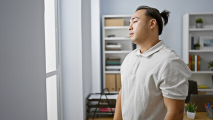 Young, handsome chinese business worker in pigtails looking through the office window with a...