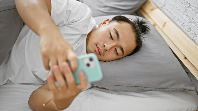 Handsome young chinese man, in relaxed fashion, lying comfortably in bed at home using smartphone in bedroom, immersed in a serious online world