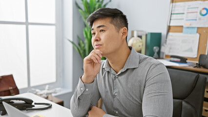 A contemplative young asian man in an office setting with a focus on business and professionalism,...