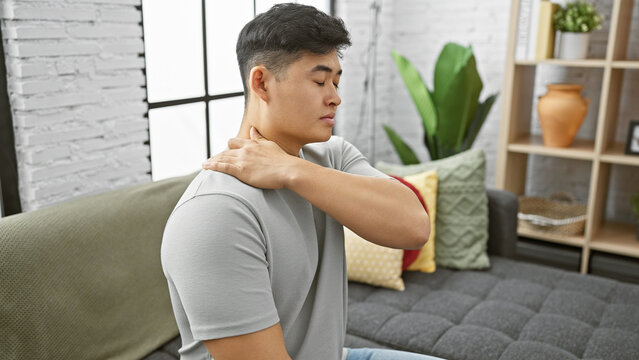 Asian Man Experiencing Neck Pain At Home, Sitting On A Gray Sofa With A Decorative Interior Background.