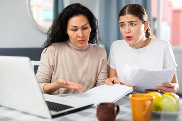 Worried pair of women holding and looking at papers and laptop at home, financial problem concept