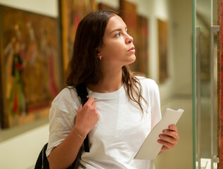 Focused girl visiting an exhibition in a museum looks with interest at the exhibit located behind...