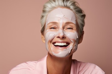 Fototapeta premium woman 50 plus, smiling with clay face mask, studio portrait on a pink background