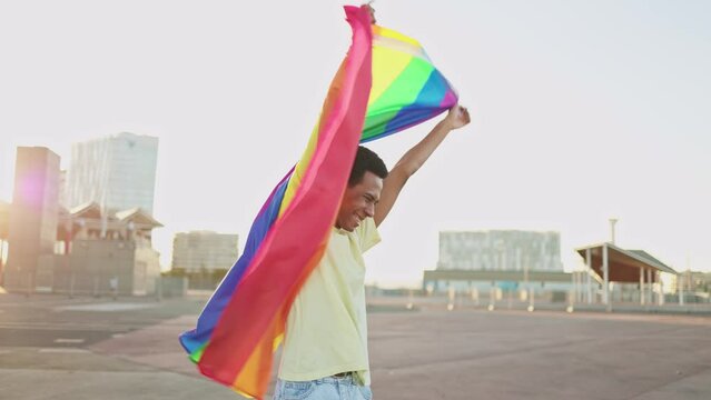 Cheerful African American non-binary person waving the LGBT rainbow flag celebrating gay pride day. Concept of LGBTQ diversity and inclusion.