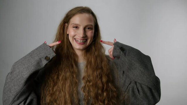 A woman points her fingers at her teeth with braces with her hands