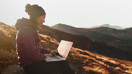 Woman working on laptop in sunset mountains. Young freelancer tourist caucasian female typing on notebook sitting on autumn grass meadow landscape. Concept of remote online work, study or business - obrazy, fototapety, plakaty
